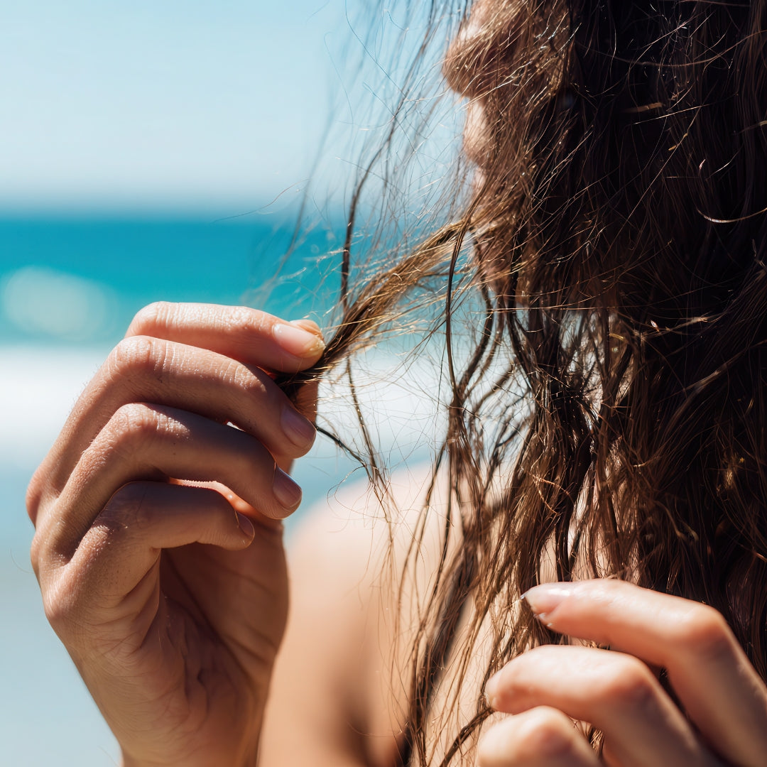 Woman on a tropical beach examining a wet hair strand to test for sun damage.